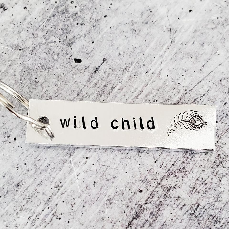 Silver rectangular metal bar keychain engraved with the words wild child and a peacock feather graphic
