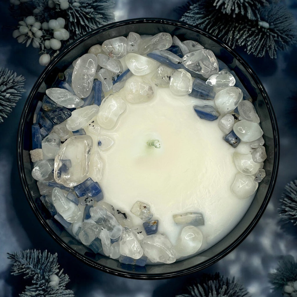 Top-down view of a Winter's Eve candle in a black jar, decorated with clear and blue crystals, set against a backdrop of frosted pine needles.