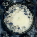 Top-down view of a Winter's Eve candle in a black jar, decorated with clear and blue crystals, set against a backdrop of frosted pine needles.