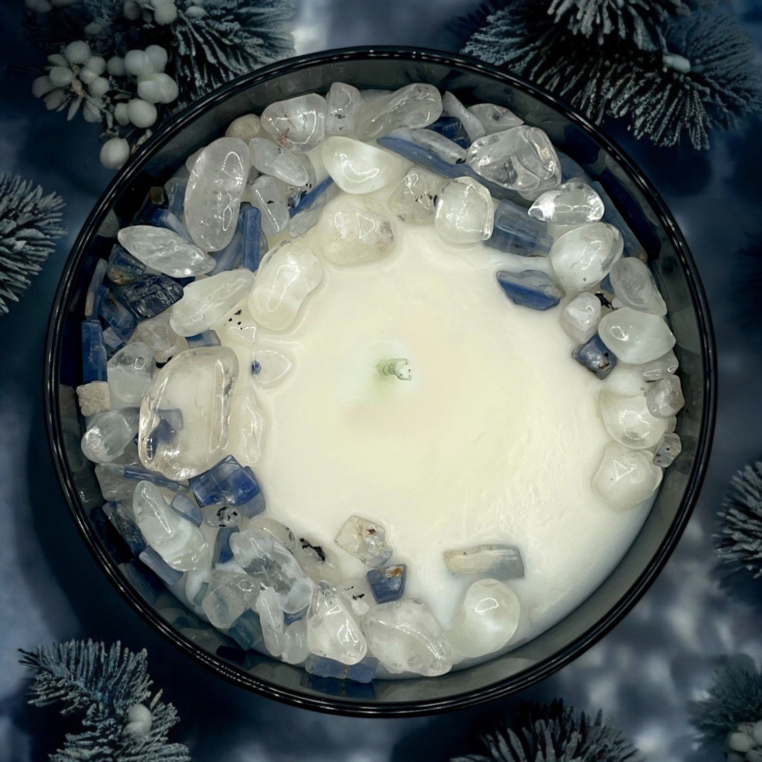Top-down view of a Winter's Eve candle in a black jar, decorated with clear and blue crystals, set against a backdrop of frosted pine needles.