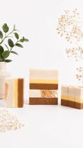 A beige and honey-yellow artisan soap bar with oatmeal flakes and goat milk, displayed in a minimalist natural setting.