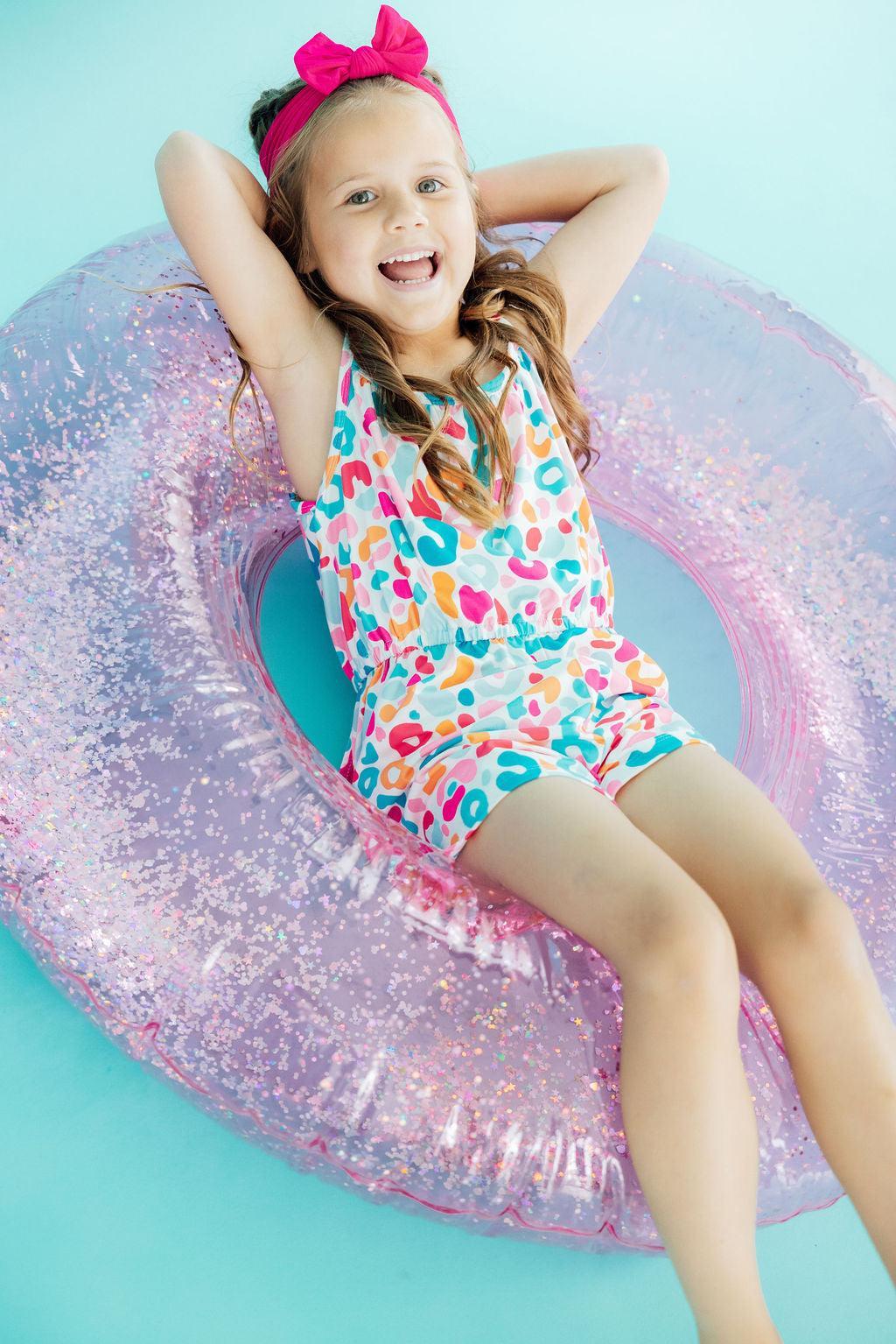 A young girl smiling while wearing a multi-colored animal print strappy play romper