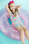 A young girl smiling while wearing a multi-colored animal print strappy play romper