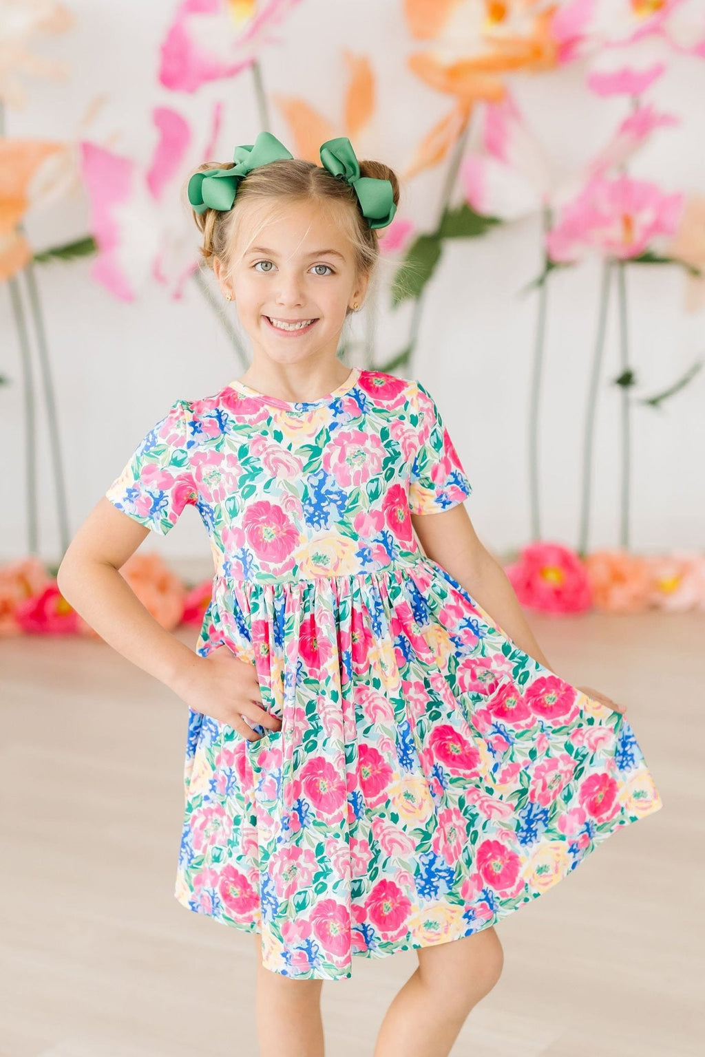 A young girl smiling wearing a bright, floral print short sleeve twirl dress with pockets.