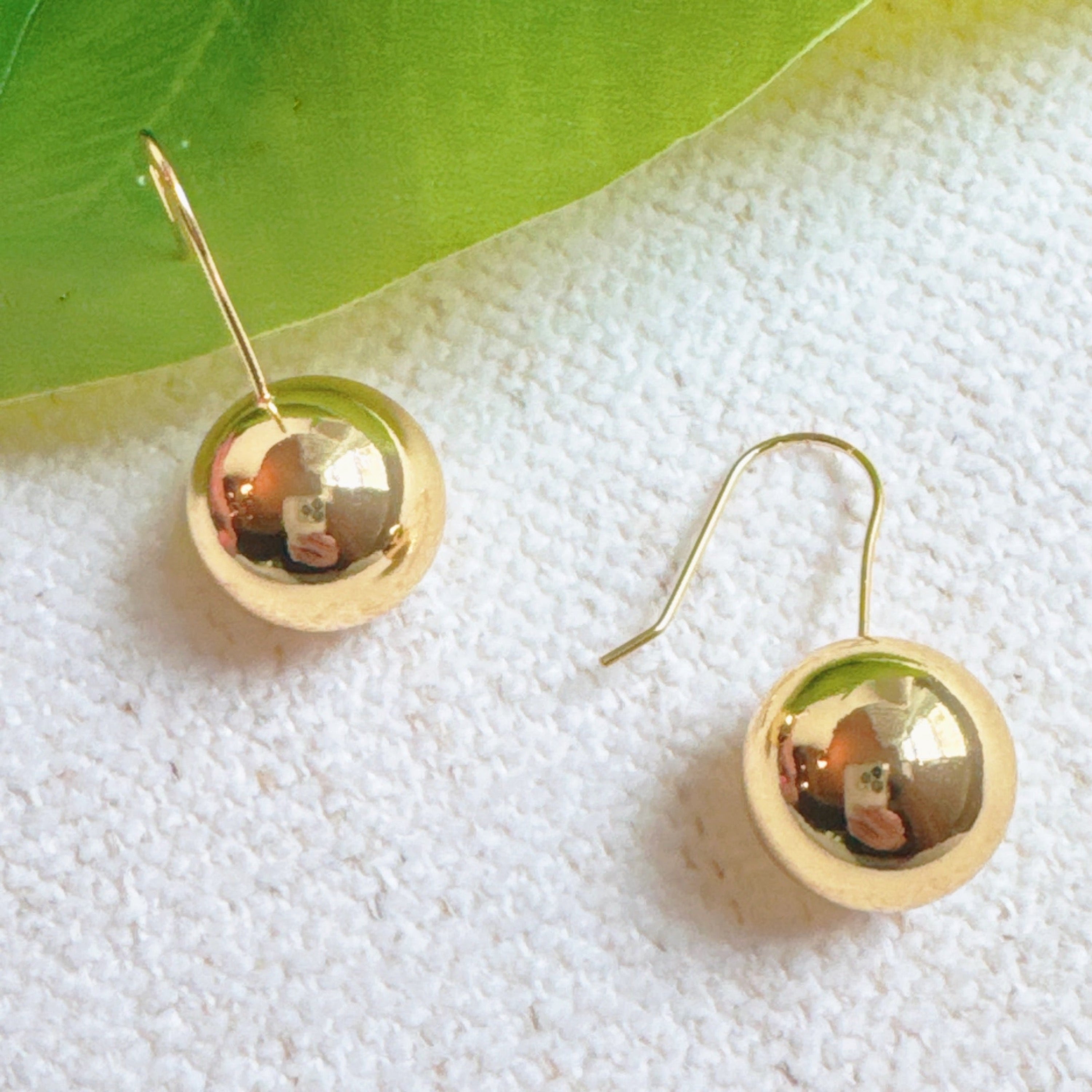 A pair of gold wire hook drop earrings with polished sphere ball pendants against a textured white background.