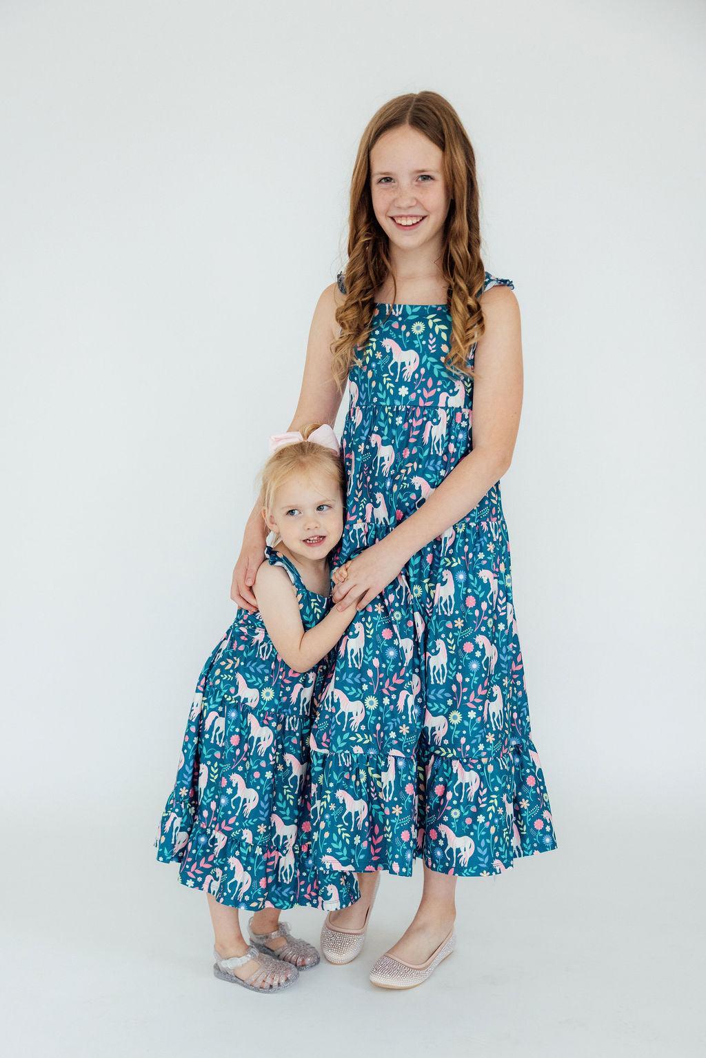 Two girls standing and holding hands wearing matching teal unicorn and floral print maxi dresses with ruffle shoulder straps and back cutouts.