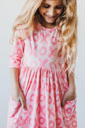 A young girl smiling while wearing a pink 3/4 sleeve dress with a red XOXO pattern and side pockets.