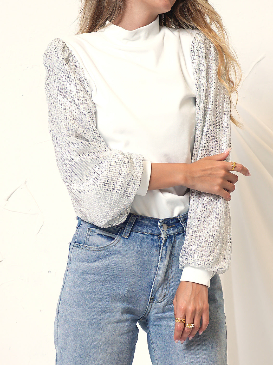 Model wearing a white mock neck long sleeve top with silver sequined balloon sleeves paired with blue jeans
