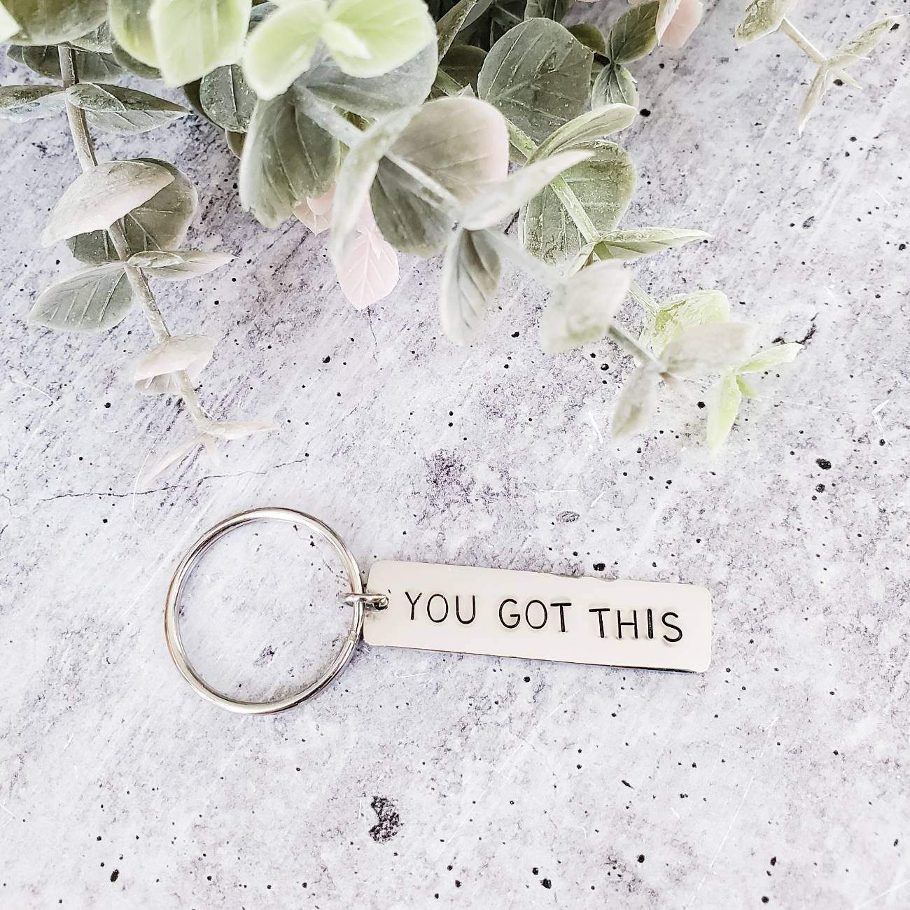 A close-up of a minimalist metal bar keychain stamped with the words YOU GOT THIS, shown in both silver and gold finishes.
