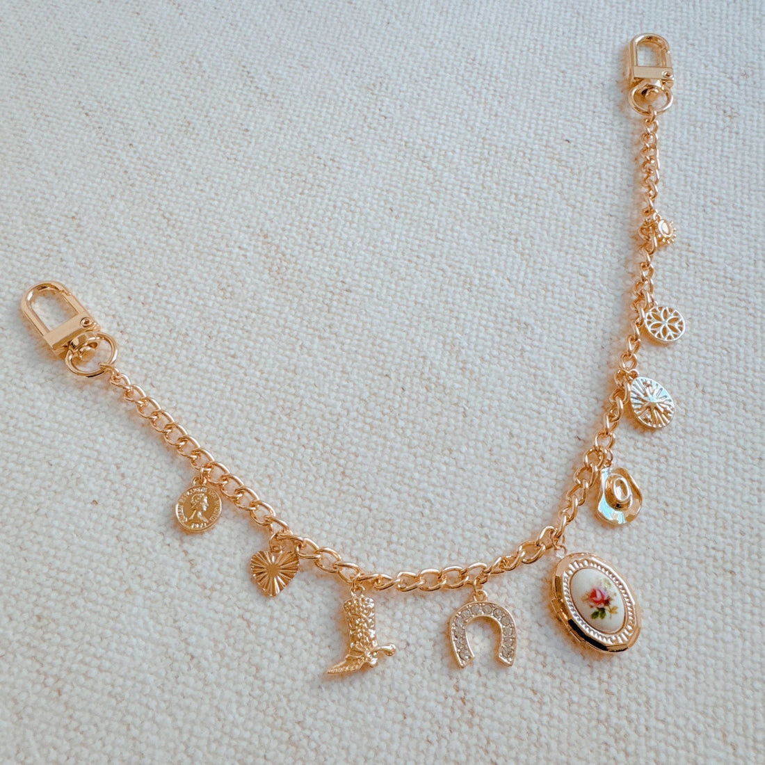 A gold-tone bag charm with western-themed charms including a cowboy boot, horseshoe, and rose locket, attached to a black handbag.