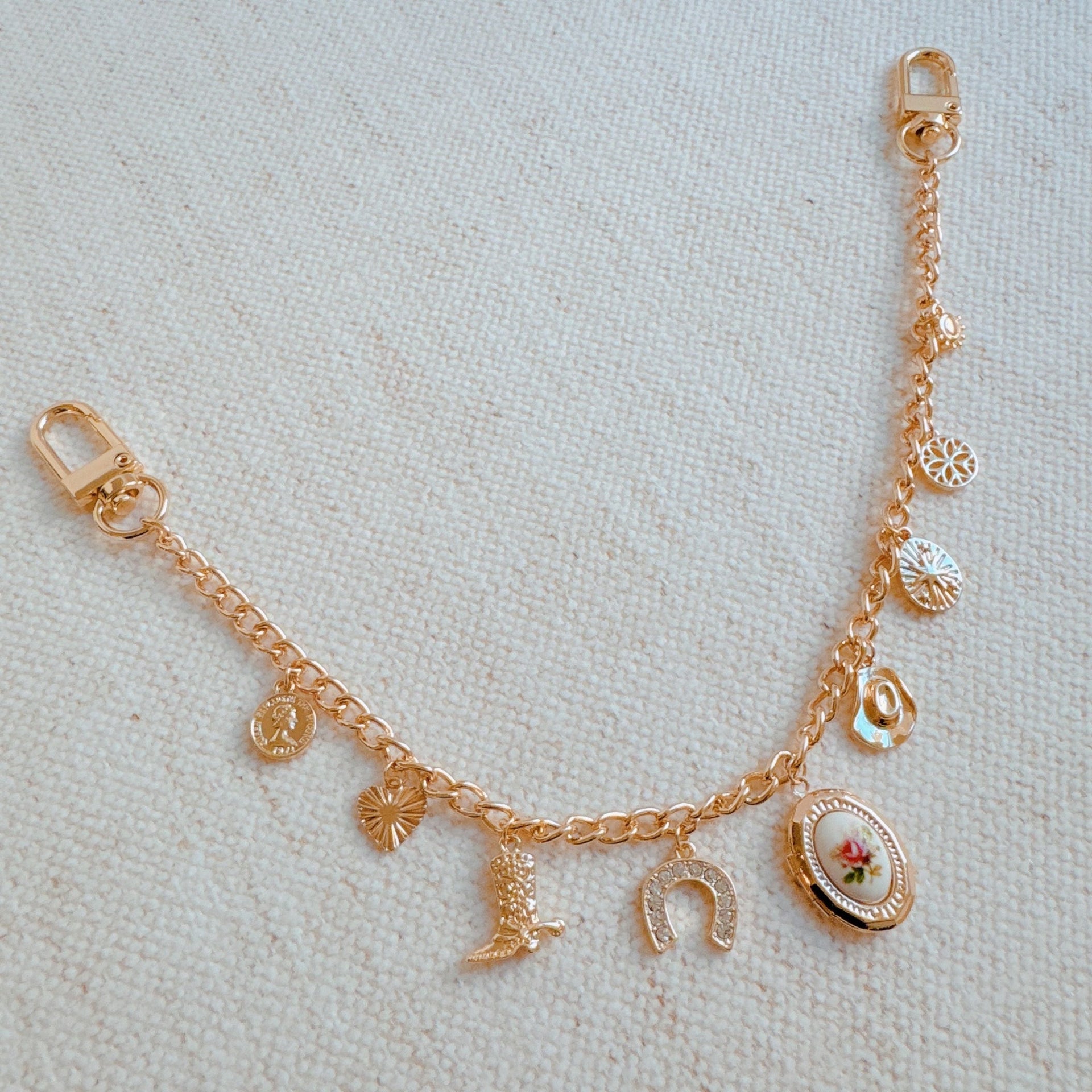A gold-tone bag charm with western-themed charms including a cowboy boot, horseshoe, and rose locket, attached to a black handbag.