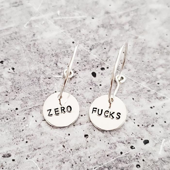A pair of hand-stamped hoop earrings with small discs reading ZERO on one and FUCKS on the other.