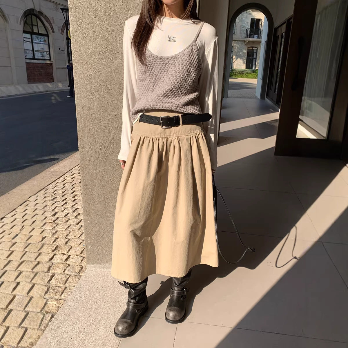A person wearing a beige A-line midi skirt with a belt, paired with a white long-sleeve top, grey knit tank, and dark boots.