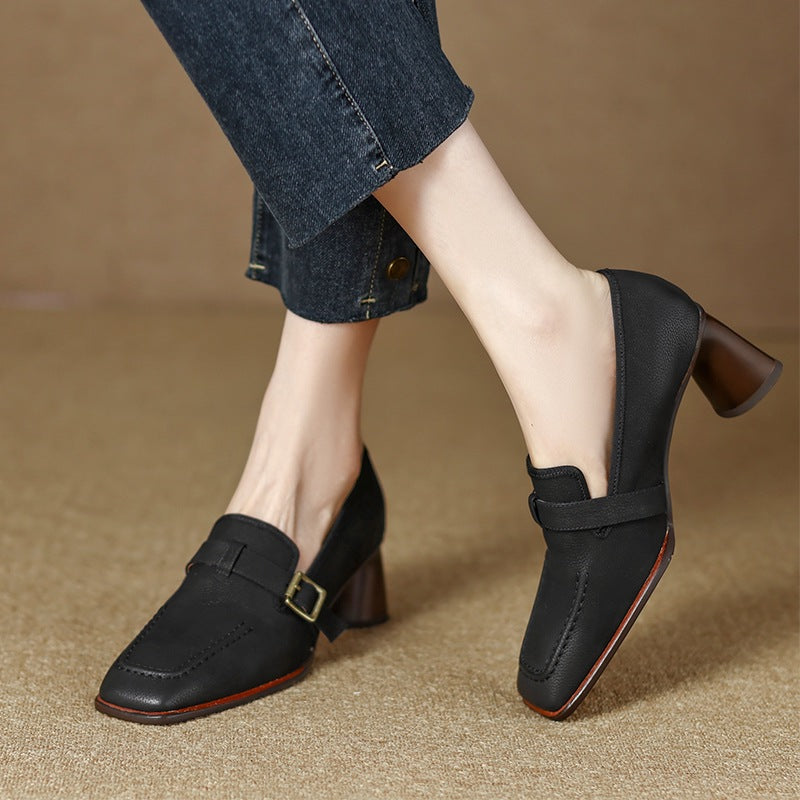 A pair of black and brown leather chunky heel shoes with a decorative buckle strap on a neutral background.