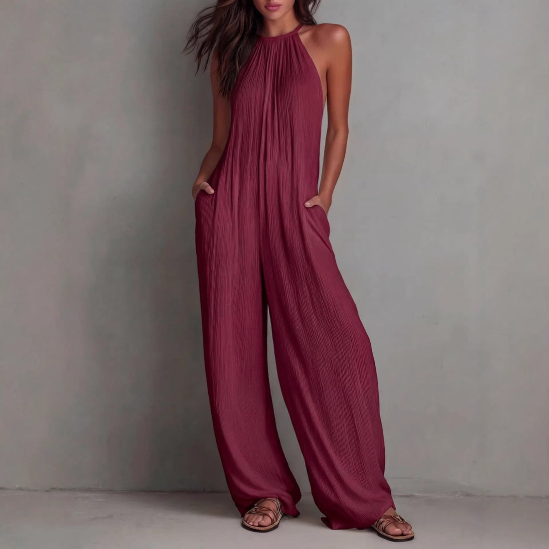 A woman wearing a sleeveless burgundy pleated wide-leg jumpsuit with an open-back design and side pockets.