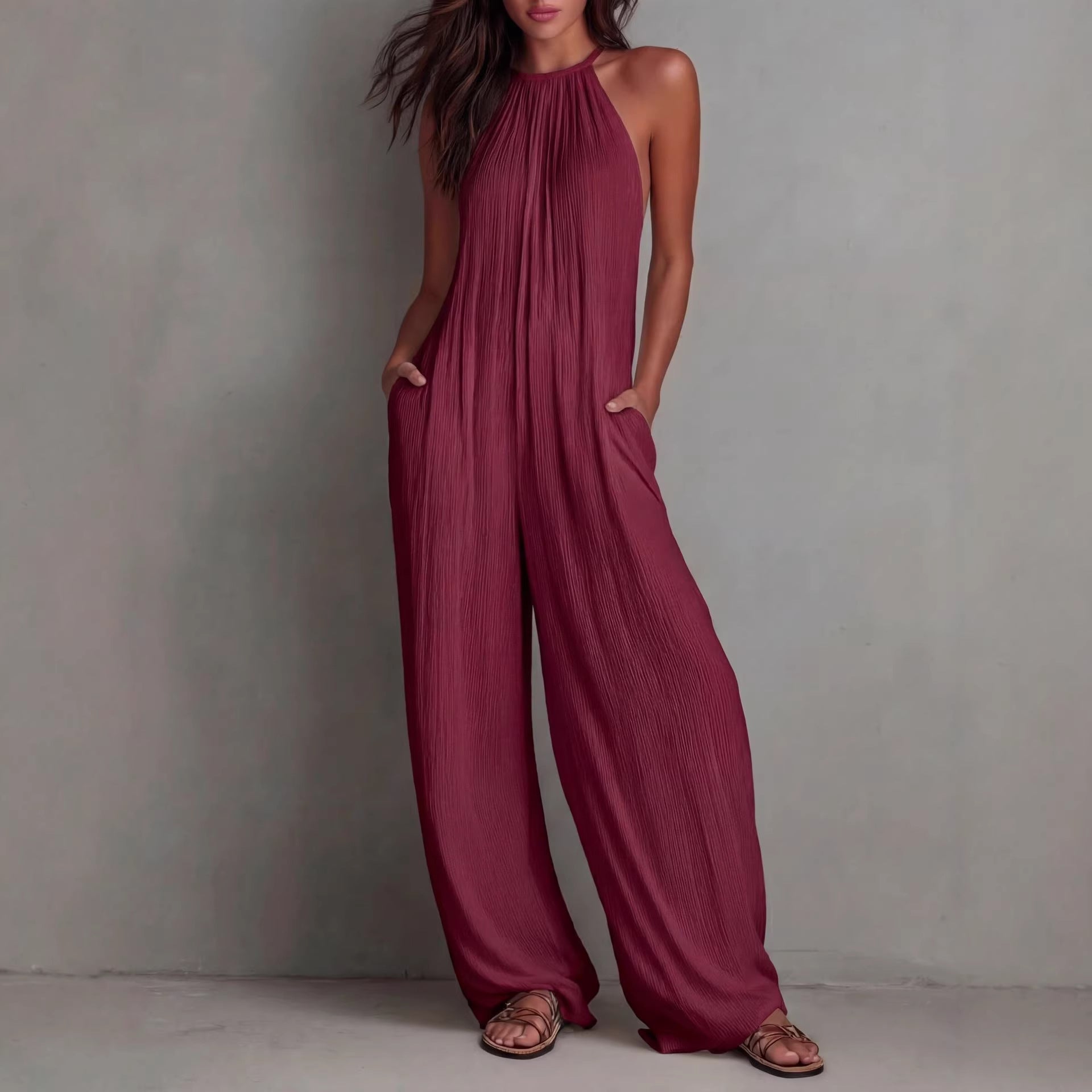A woman wearing a sleeveless burgundy pleated wide-leg jumpsuit with an open-back design and side pockets.