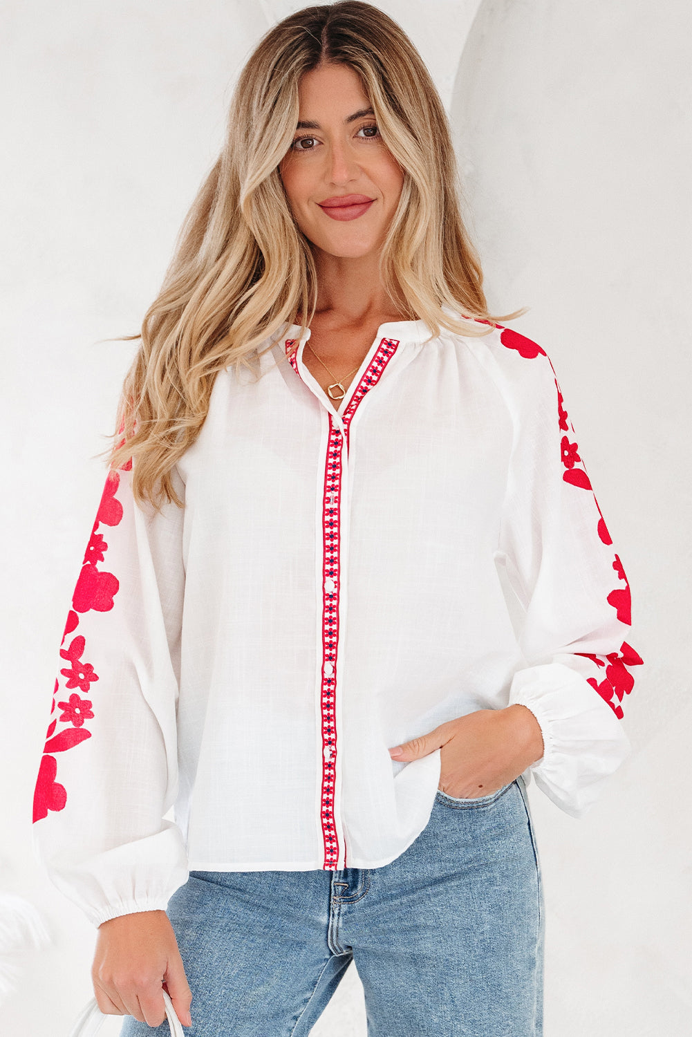 A woman wearing a white long-sleeve blouse with bold red floral embroidery on the sleeves and front placket.