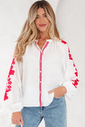 A woman wearing a white long-sleeve blouse with bold red floral embroidery on the sleeves and front placket.