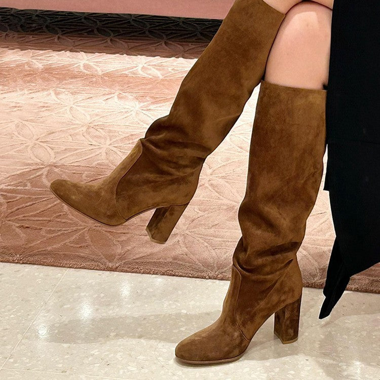 A pair of brown suede knee-high boots with a pointed toe and chunky block heel, featuring a pleated shaft detail.