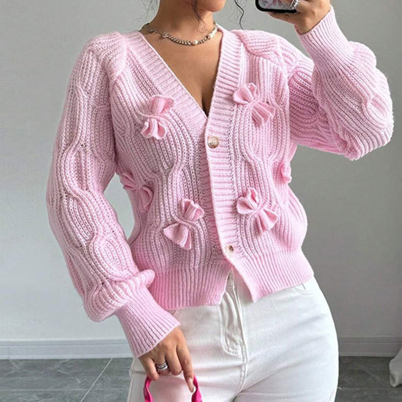 A woman wearing a light pink, cropped, V-neck cardigan featuring multiple small bow details and a ribbed knit texture.