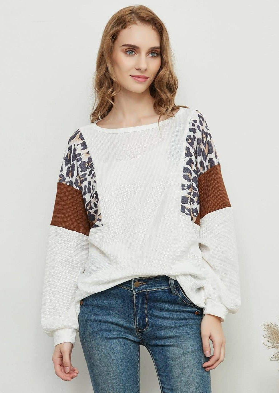 A woman wearing a white waffle knit pullover sweater with brown accents and leopard print sleeves, paired with blue jeans.
