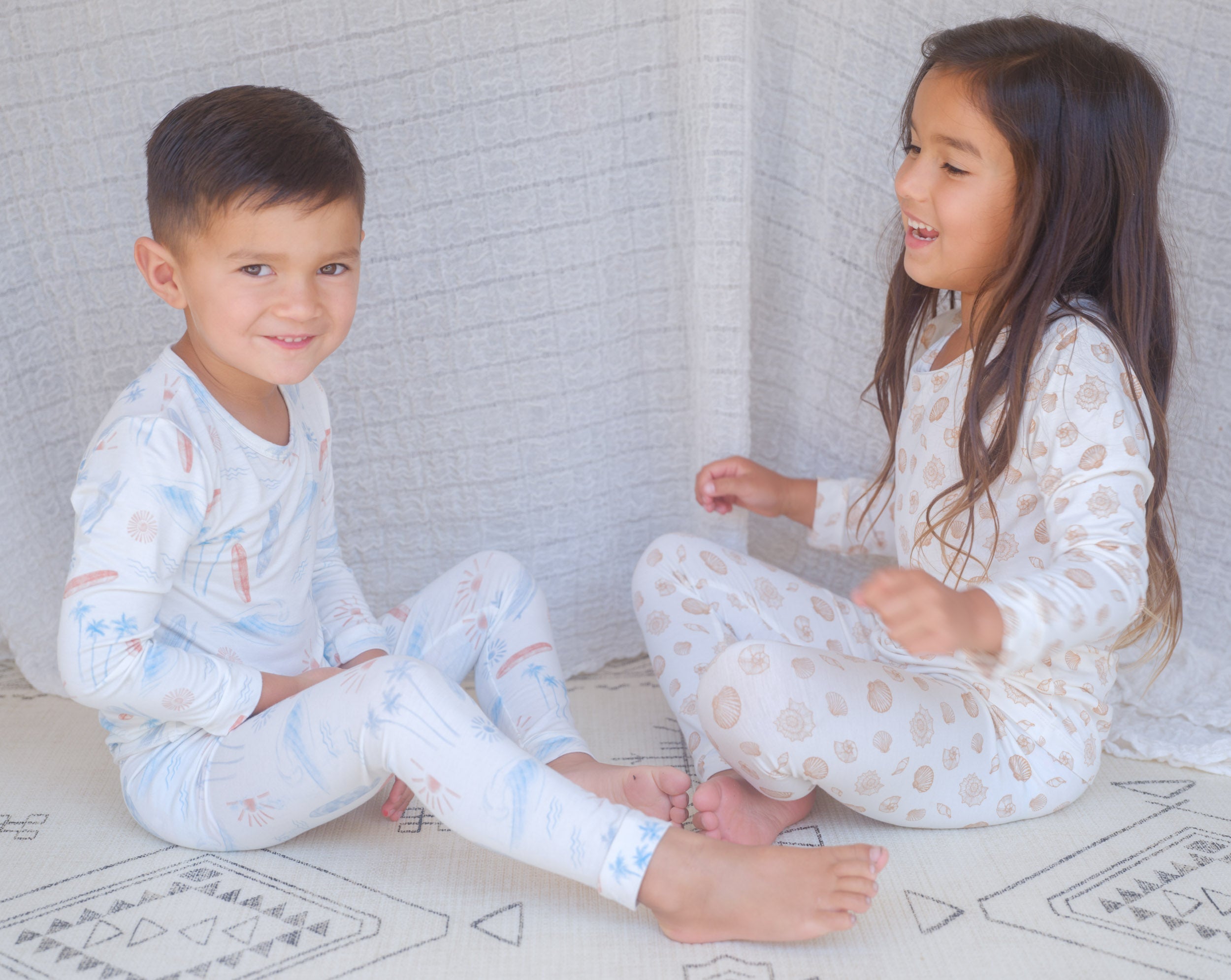 Two children in matching pajamas sitting on a rug against a white brick wall.