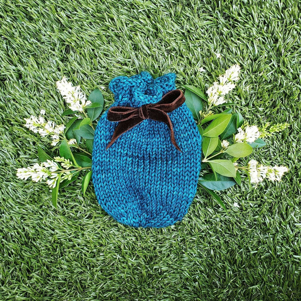 Handmade teal knit avocado ripening pouch with a dark brown tie closure sitting on a wooden surface.