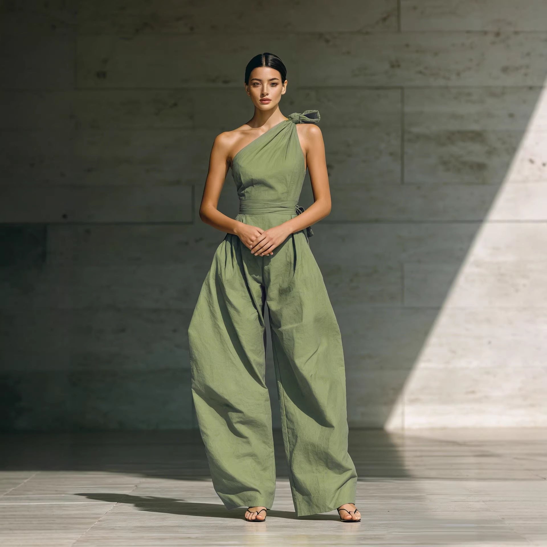 A woman wearing a sage green one-shoulder jumpsuit with a wide-leg cut and a matching waist belt.