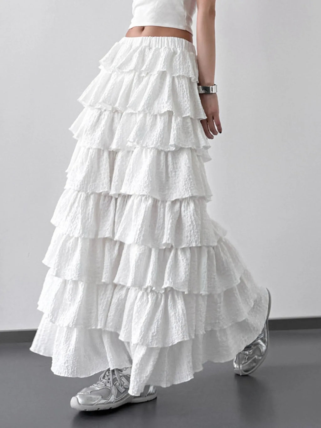A woman wearing a white ruffled tiered maxi skirt paired with silver sneakers against a neutral background.