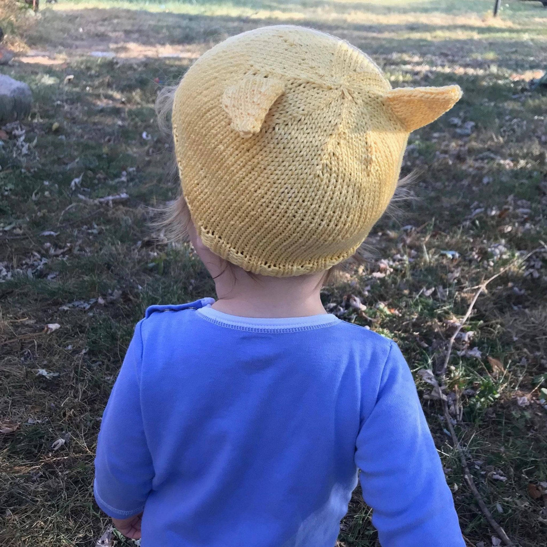 A bright yellow handmade knit baby beanie with adorable kitten ears, displayed in a sunny, whimsical lifestyle setting.