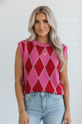 Woman wearing a red and pink argyle sleeveless top with light blue jeans against a white brick wall.