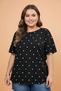 A flat lay of a black short sleeve t-shirt featuring a white polka dot pattern and a decorative scalloped hem.