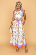 A woman wearing a white floral maxi dress with orange and lavender trim and a removable belt against a beige background