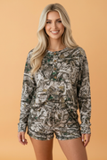 Woman wearing a matching camo long sleeve henley top and elastic waist shorts loungewear set in olive green, brown, and tan.