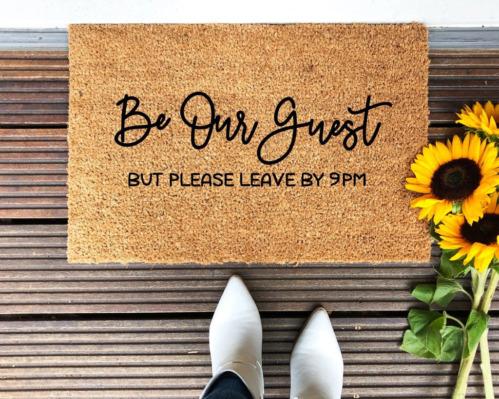 A brown natural coir doormat with black text reading Be Our Guest, But Please Leave By 9 on a porch.
