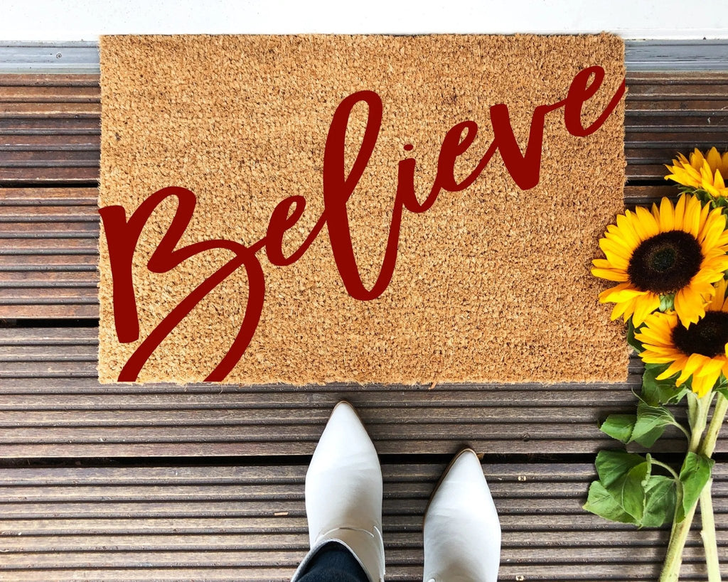 Rustic Believe holiday doormat with red script on natural coir at an outdoor entryway.