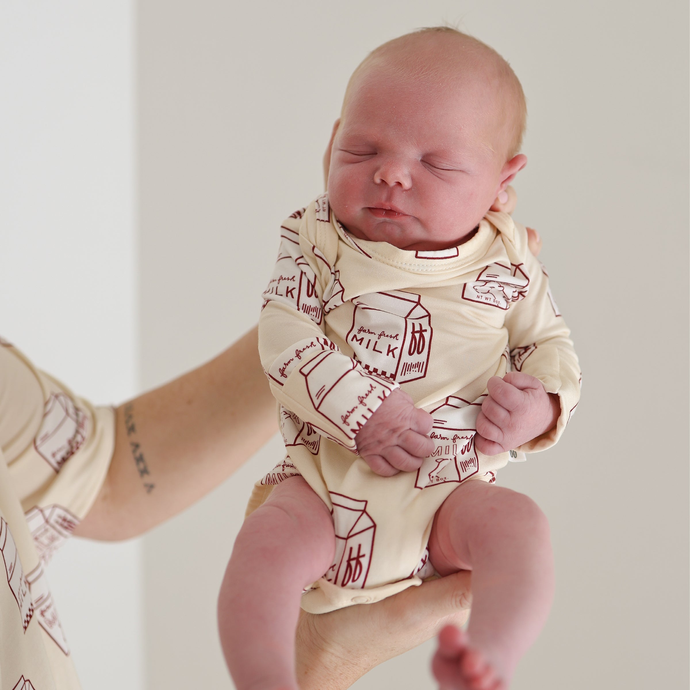 A newborn baby wearing a cream-colored long-sleeve snapsuit with a red milk carton print.
