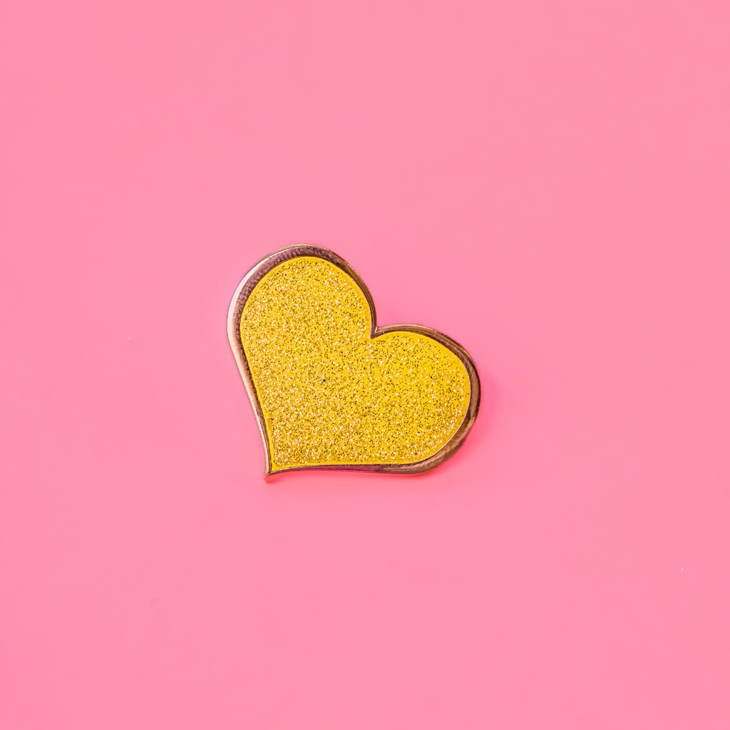 A close-up of a yellow heart-shaped enamel pin with a glittering surface against a solid pink background.