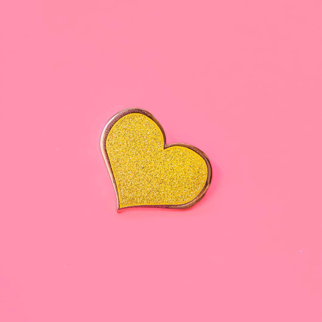 A close-up of a yellow heart-shaped enamel pin with a glittering surface against a solid pink background.