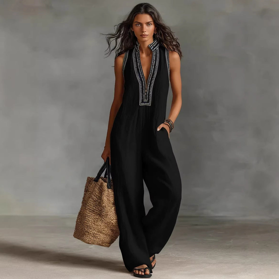 A woman wearing a black sleeveless linen-blend jumpsuit with a loose waist and full-length pants, paired with a large straw tote bag.