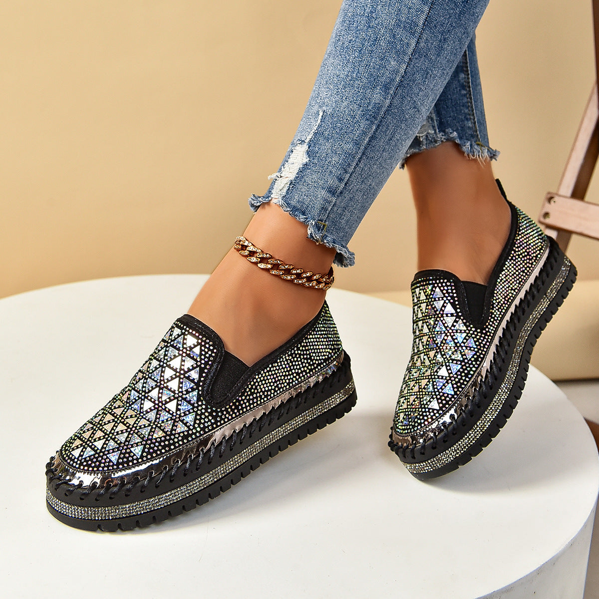 Close up of a pair of women's black slip-on sneakers with rhinestone geometric patterns and a thick sole worn with ripped jeans.