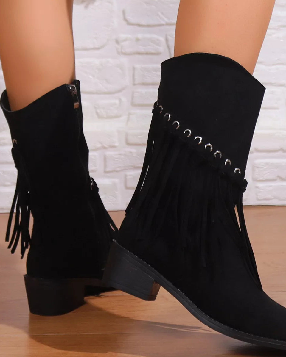 Black suede boots with fringe details worn by a person against a white brick wall.