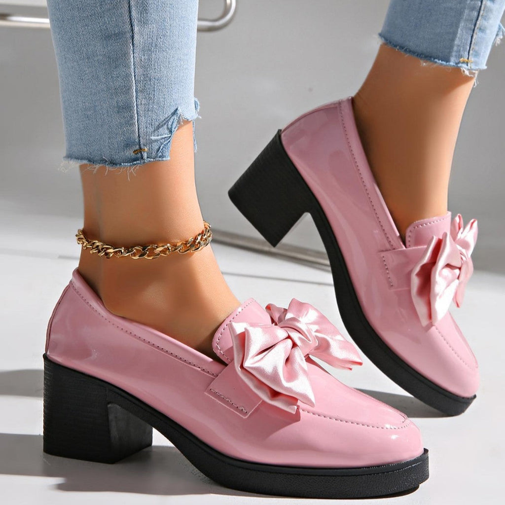 A pair of glossy pink patent block heel pumps with a large fabric bow on the toe, worn with light wash denim jeans.