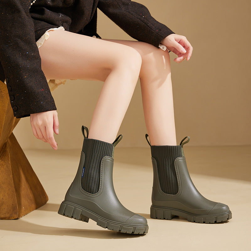 A pair of olive green waterproof ankle rain boots with a lug sole and ribbed knit upper worn by a model.