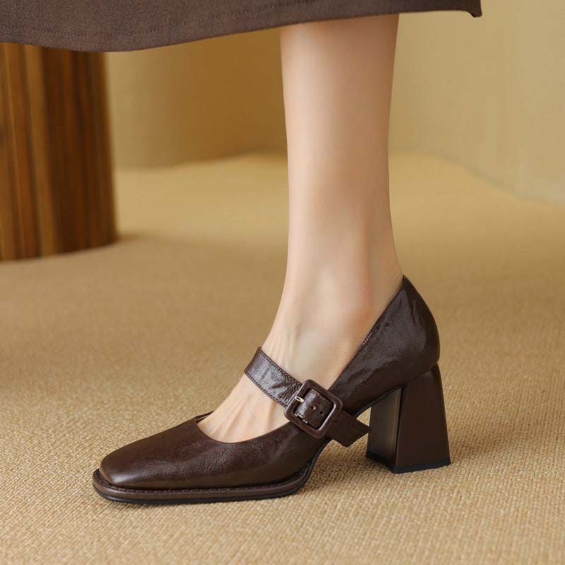 A woman wearing black leather square-toe Mary Jane heels with a chunky block heel and buckled strap.