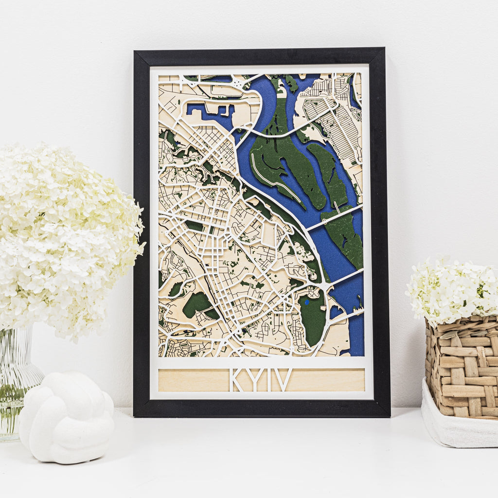 3D wooden city map of Kyiv with blue and green geometric details in a black frame hanging on a wall.