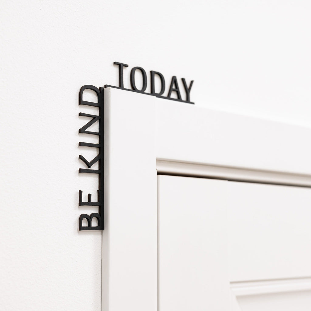 Minimalist black wooden 'Be Kind Today' wall quote installed in a modern room corner.