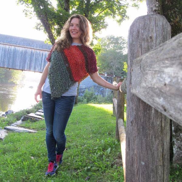 Woman wearing an oversized striped knit poncho in olive, orange, maroon, and mustard colors in a rustic setting.