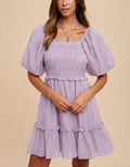 A lilac mini dress featuring a smocked bodice, square neckline, and tiered skirt with short bubble sleeves.