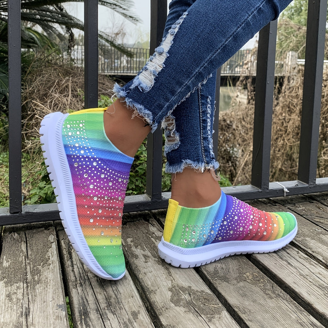 A person wearing vibrant rainbow-colored mesh slip-on sneakers with rhinestone embellishments on a wooden walkway.